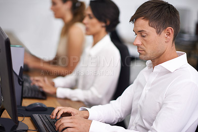 Buy stock photo Shot of a handsome young office worker at his computer with his colleagues in the background