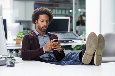 Buy stock photo Shot of a young businessman sending a text message with his feet on a desk