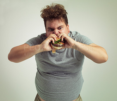 Buy stock photo Takeaway, weight gain and burger for man in studio with fastfood for unhealthy eating, plus size and greed. Young person with meal in mouth and fat stomach for hunger, meat and sauce for bite indoor