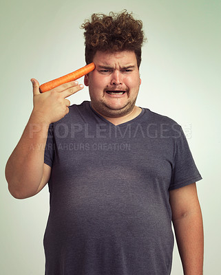 Buy stock photo Depression, carrot and hand gun with plus size man in studio on gray background for diet, health or nutrition. Sad, unhappy and vegetable to head of young person in conflict with vegetarian food