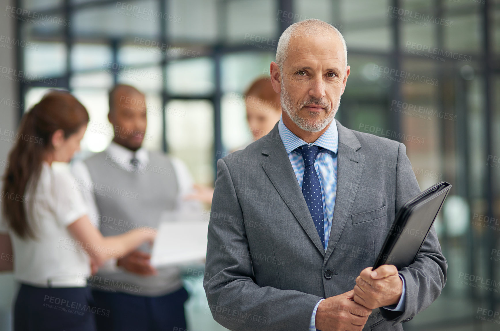 Buy stock photo Portrait of a mature businessman standing in the office with his colleagues in the background