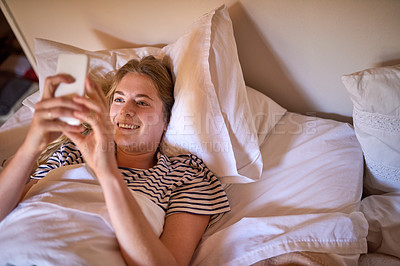 Buy stock photo Shot of a happy young woman using her cellphone while lying in bed