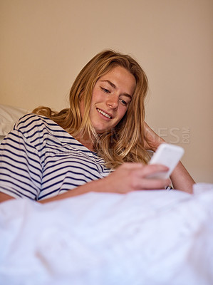 Buy stock photo Shot of a happy young woman using her cellphone while lying in bed