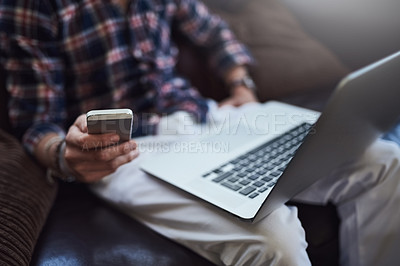 Buy stock photo High angle shot of an unrecognizable man using his laptop and cellphone while sitting on the sofa at home