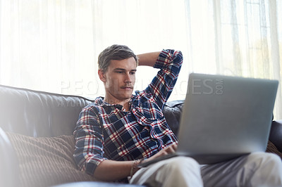 Buy stock photo Shot of a handsome young man using his laptop while sitting on the sofa at home