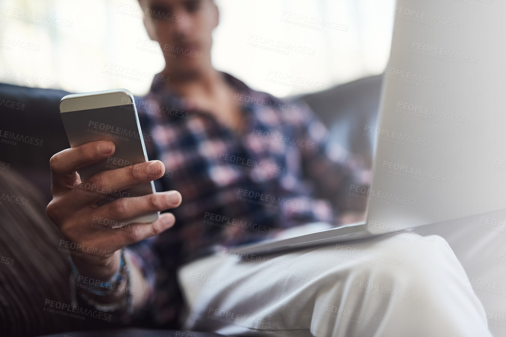 Buy stock photo Shot of a young man using his laptop and cellphone while sitting on the sofa at home