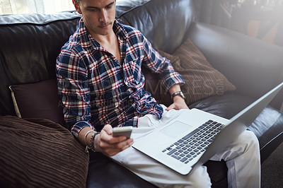 Buy stock photo High angle shot of a handsome young man using his laptop and cellphone while sitting on the sofa at home