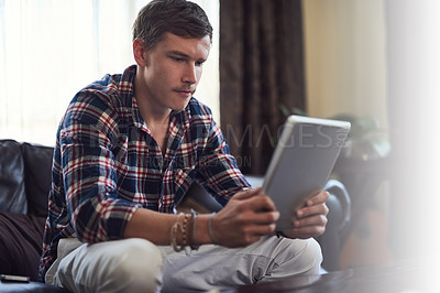 Buy stock photo Shot of a handsome young man using his tablet while sitting on the sofa at home