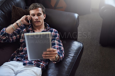 Buy stock photo High angle shot of a handsome young man using his tablet and cellphone while sitting on the sofa at home