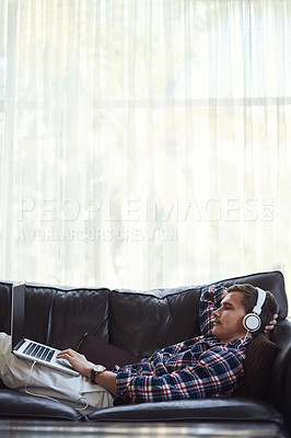 Buy stock photo Shot of a handsome young man using his laptop to listen to music while chilling on the sofa at home