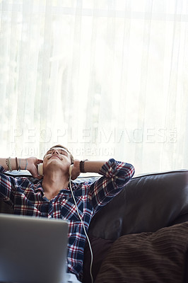Buy stock photo Shot of a handsome young man using his laptop to listen to music while sitting on the sofa at home