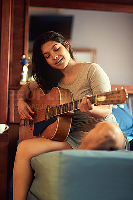 Buy stock photo Shot of a happy young woman playing her guitar while sitting on her bed
