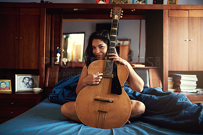 Buy stock photo Portrait of a happy young woman holding her guitar in front of her while sitting on her bed