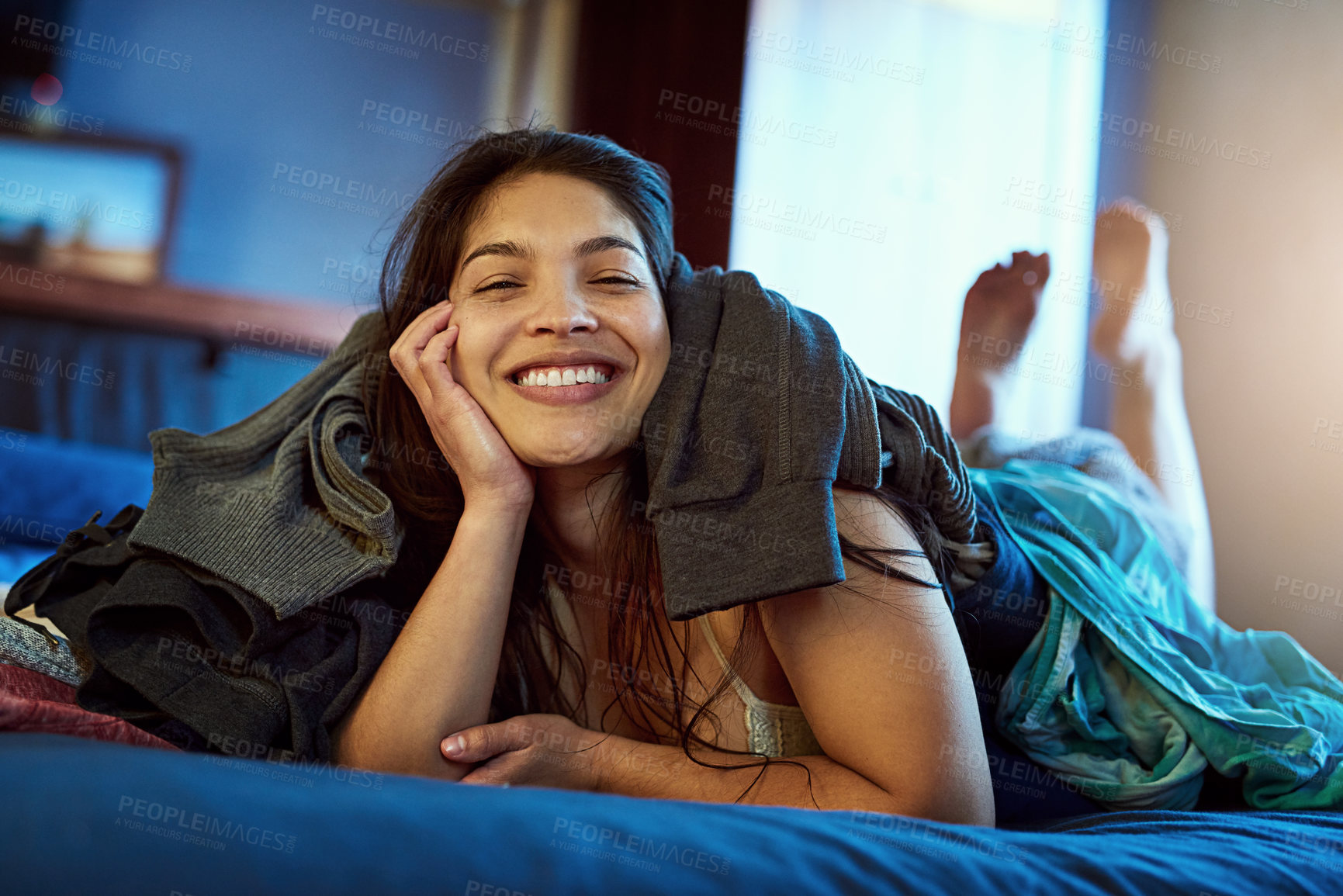 Buy stock photo Portrait of a happy young woman lying under a pile of clothes on her bed