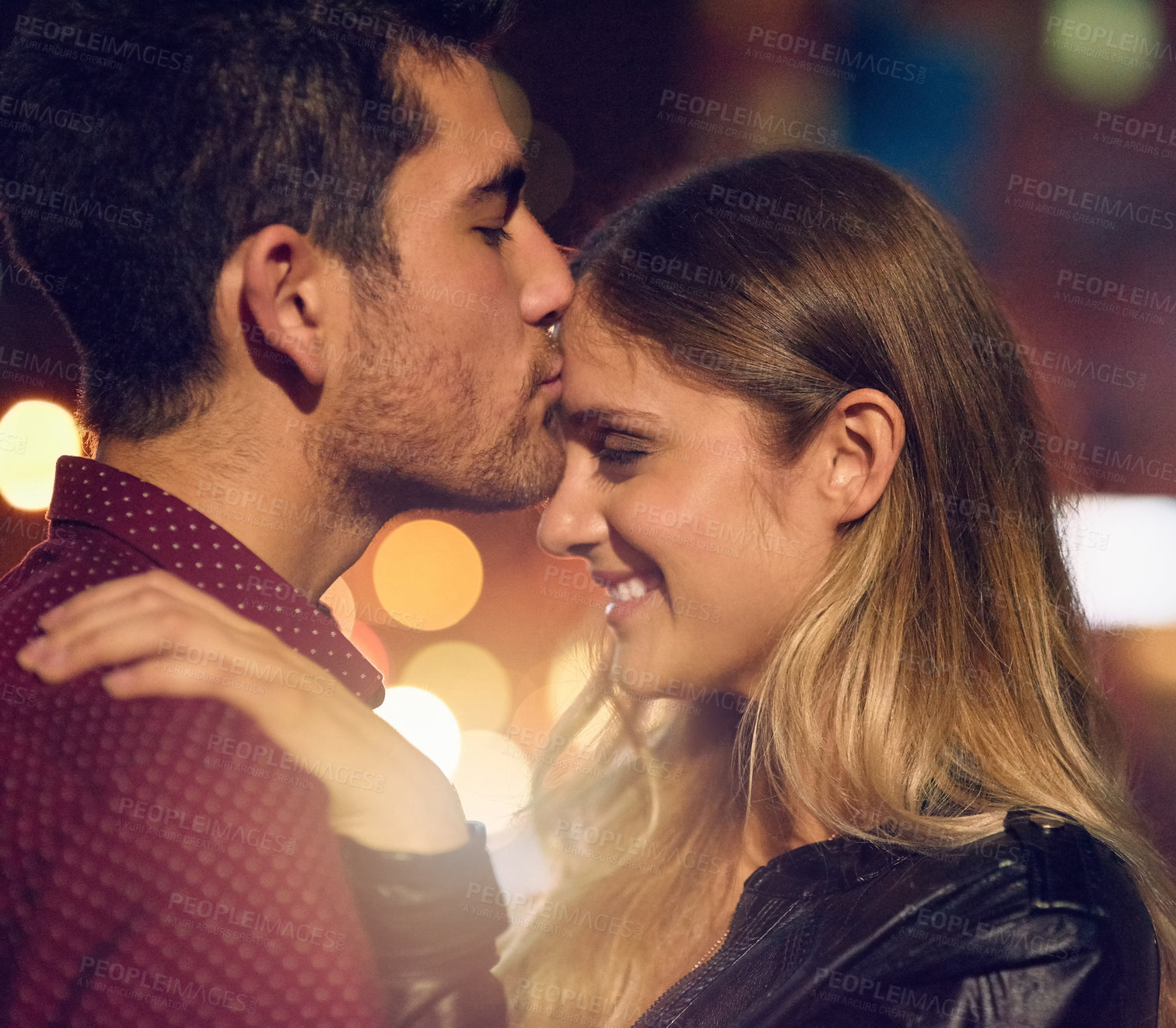 Buy stock photo Shot of a young man kissing his girlfriend on her forehead at night outside