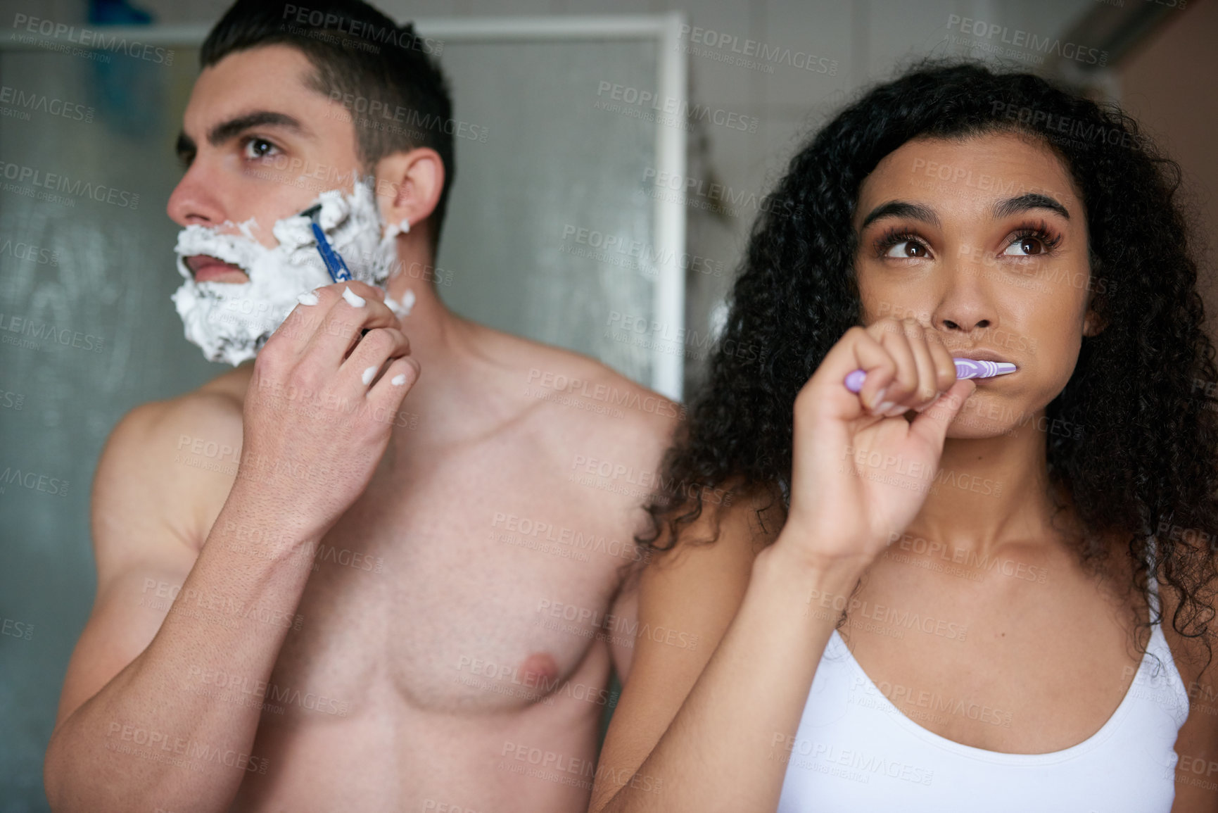 Buy stock photo Shot of an attractive young woman brushing her teeth while her boyfriend shaves in the bathroom