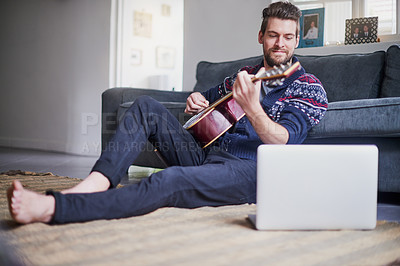 Buy stock photo Shot of a handsome young man playing a guitar while watching an online tutorial on his laptop
