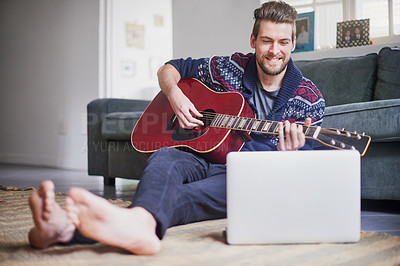 Buy stock photo Shot of a handsome young man playing a guitar while watching an online tutorial on his laptop
