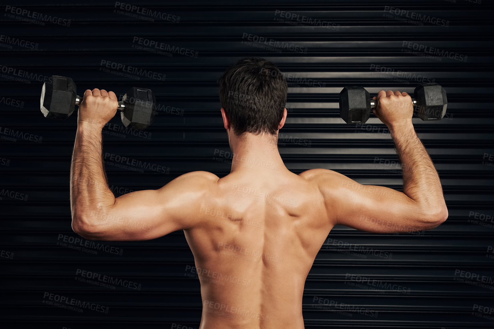 Buy stock photo Rearview shot of a shirtless and well built man lifting dumbbells warming up against a dark background