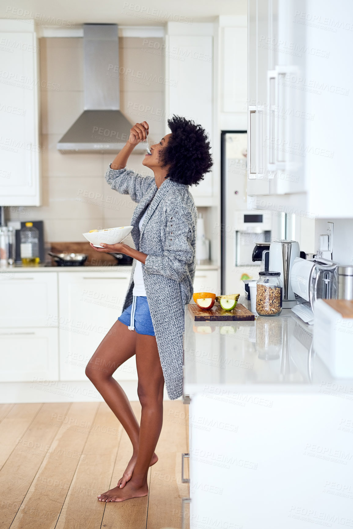 Buy stock photo Shot of a relaxed young woman eating breakfast while standing in her kitchen at home
