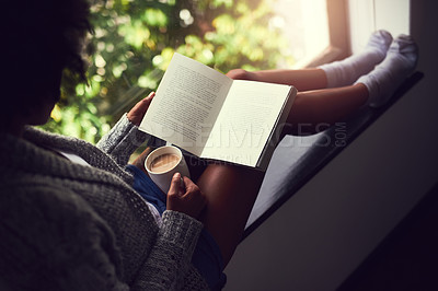 Buy stock photo Shot of an unidentifiable young woman reading a book while enjoying a cup of coffee at home