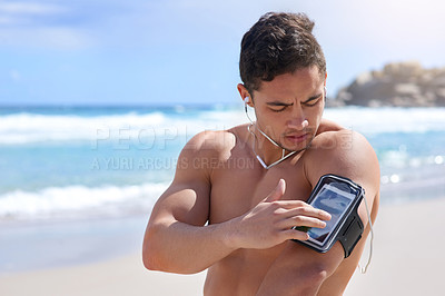 Buy stock photo Shot of a handsome young man setting up his workout playlist on the beach