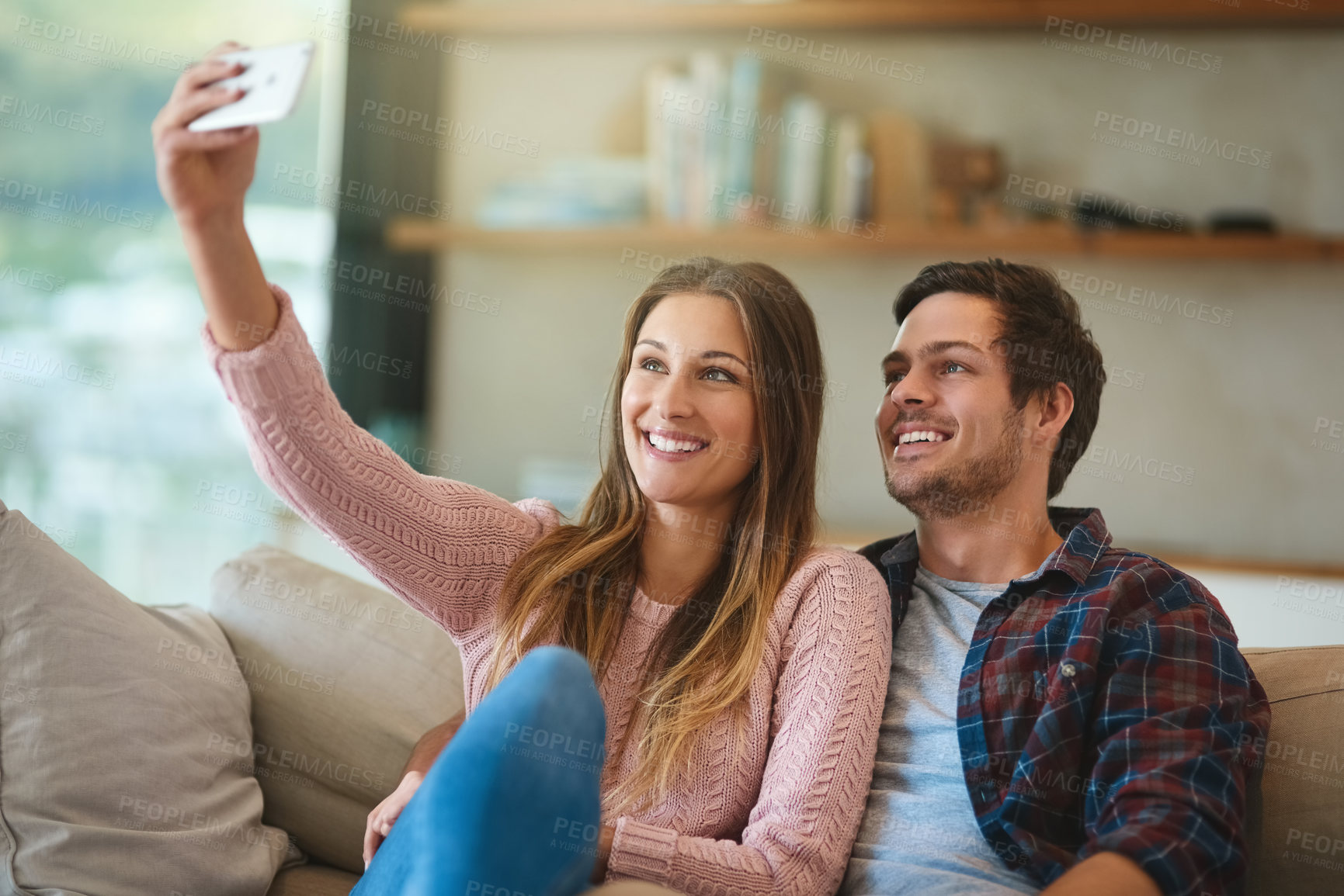 Buy stock photo Shot of a happy young couple taking a selfie with a smartphone while sitting on their couch at home