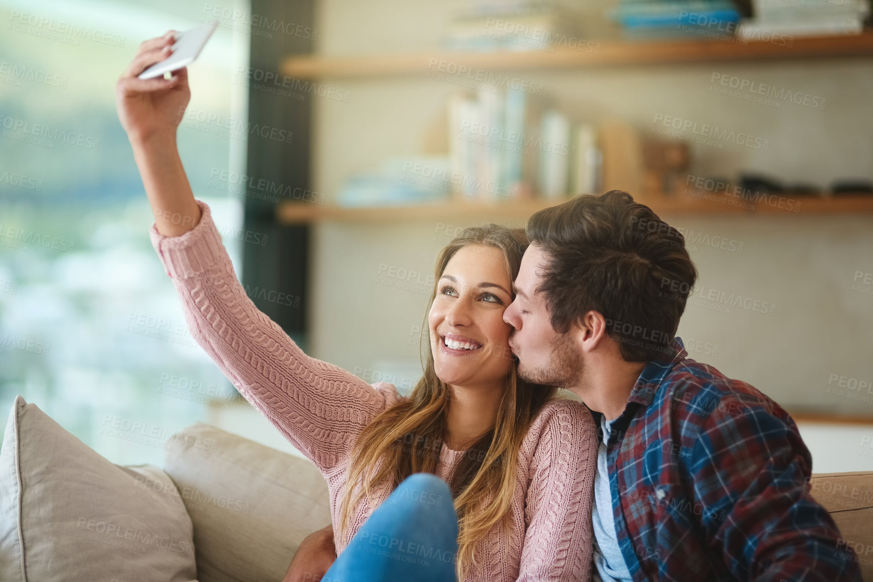 Buy stock photo Shot of a happy young couple taking a selfie with a smartphone while sitting on their couch at home