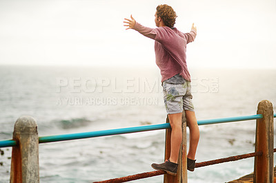 Buy stock photo Shot of a young man standing on a railing by the ocean with his arms raised in the air