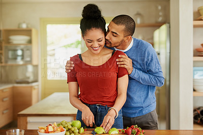 Buy stock photo Shot of a young man kissing his wife while she prepares a healthy snack at home