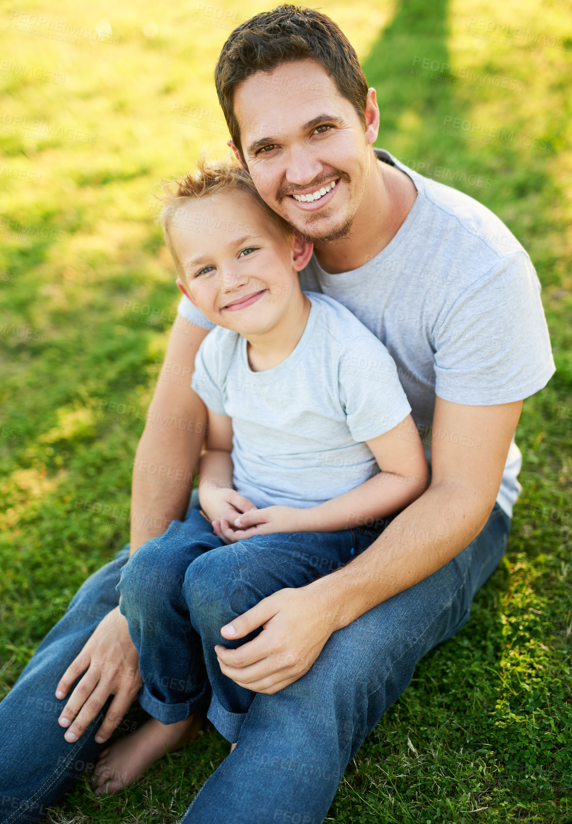 Buy stock photo Portrait of a smiling father and his little son sitting on the grass in a park