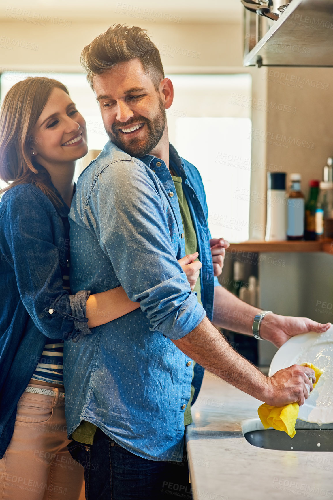 Buy stock photo Portrait of a young happy woman hugging her husband while he washes the dishes at home