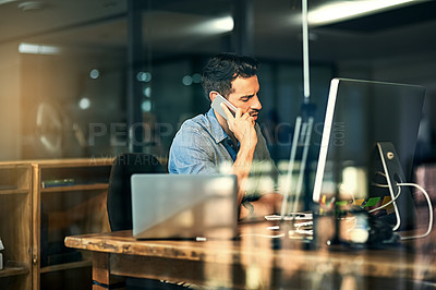 Buy stock photo Shot of a young businessman talking on his phone and using a computer during a late night at work