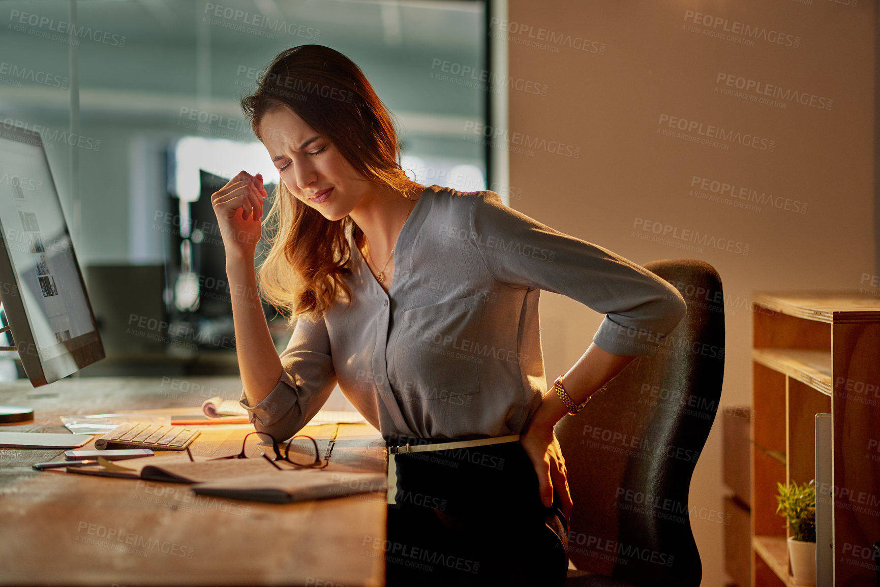 Buy stock photo Shot of an attractive young businesswoman holding her back in pain while working late in the office