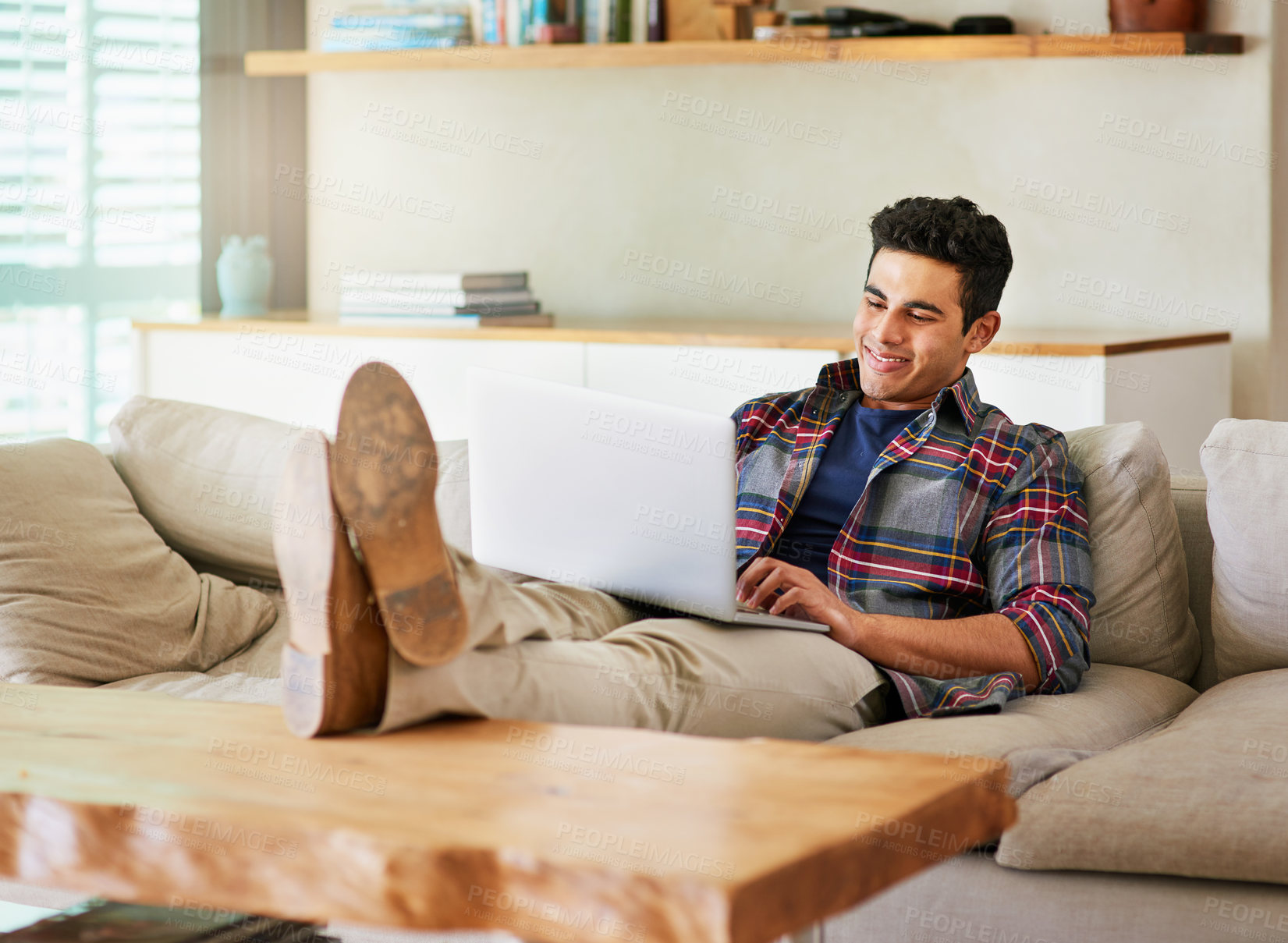 Buy stock photo Shot of a handsome young man using his laptop while relaxing on the sofa at home