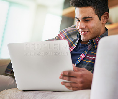 Buy stock photo Shot of a handsome young man using his laptop while relaxing on the sofa at home