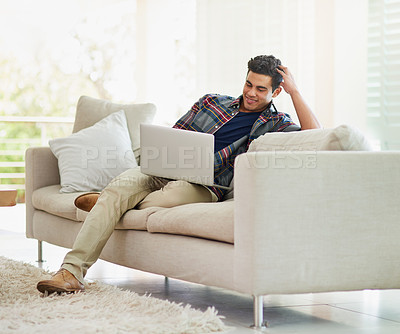 Buy stock photo Shot of a handsome young man using his laptop while relaxing on the sofa at home
