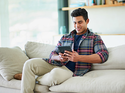 Buy stock photo Shot of a handsome young man using his tablet while relaxing on the sofa at home