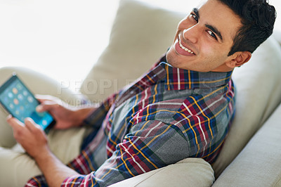 Buy stock photo Portrait of a handsome young man using his tablet while relaxing on the sofa at home
