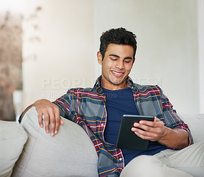 Buy stock photo Shot of a handsome young man using his tablet while relaxing on the sofa at home