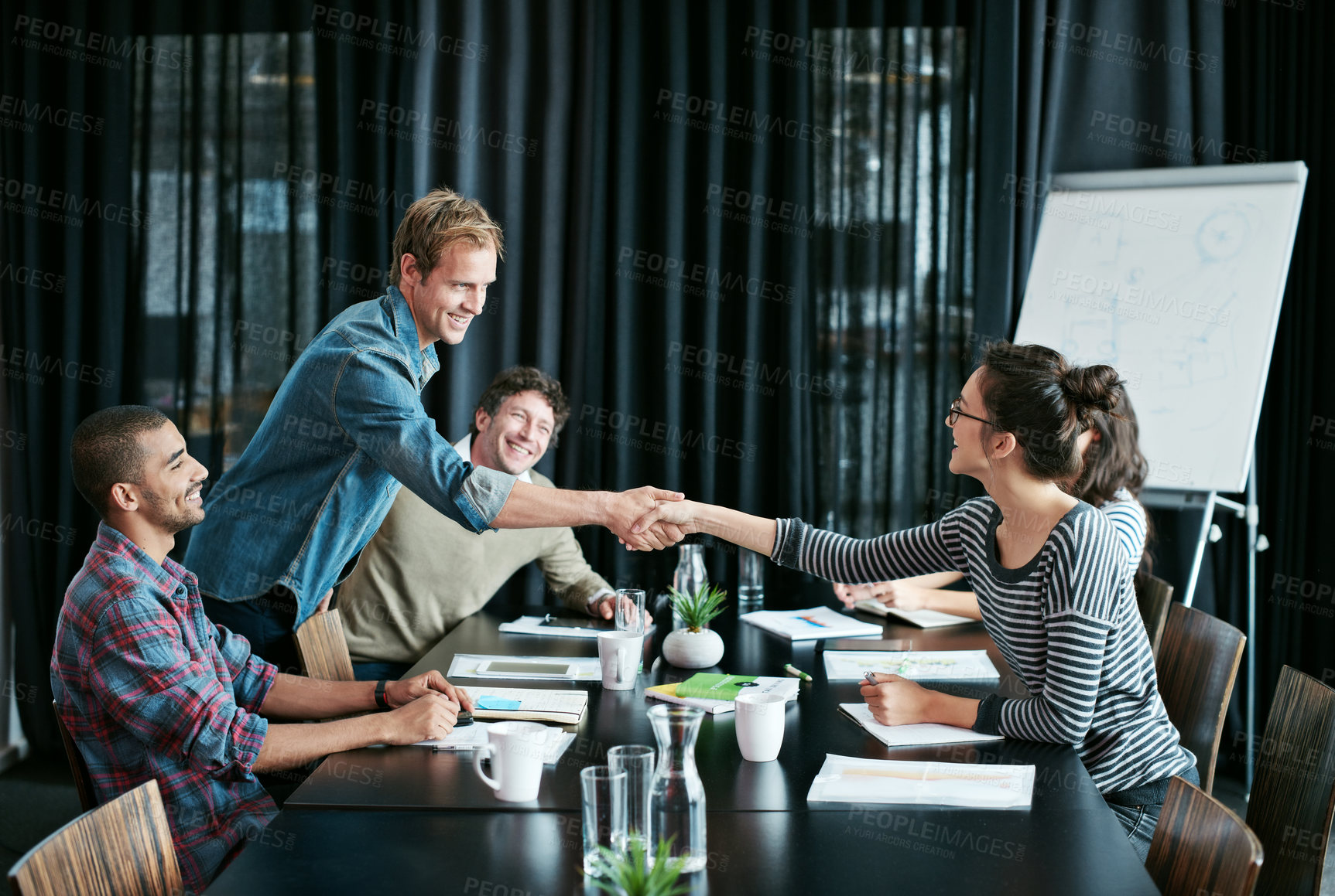 Buy stock photo Shot of two colleagues shaking hands in a boardroom while coworkers look on