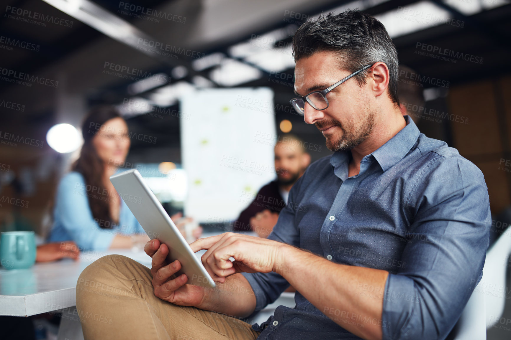 Buy stock photo Shot of a man sitting at a table in an office using a digital tablet with colleagues working in the background