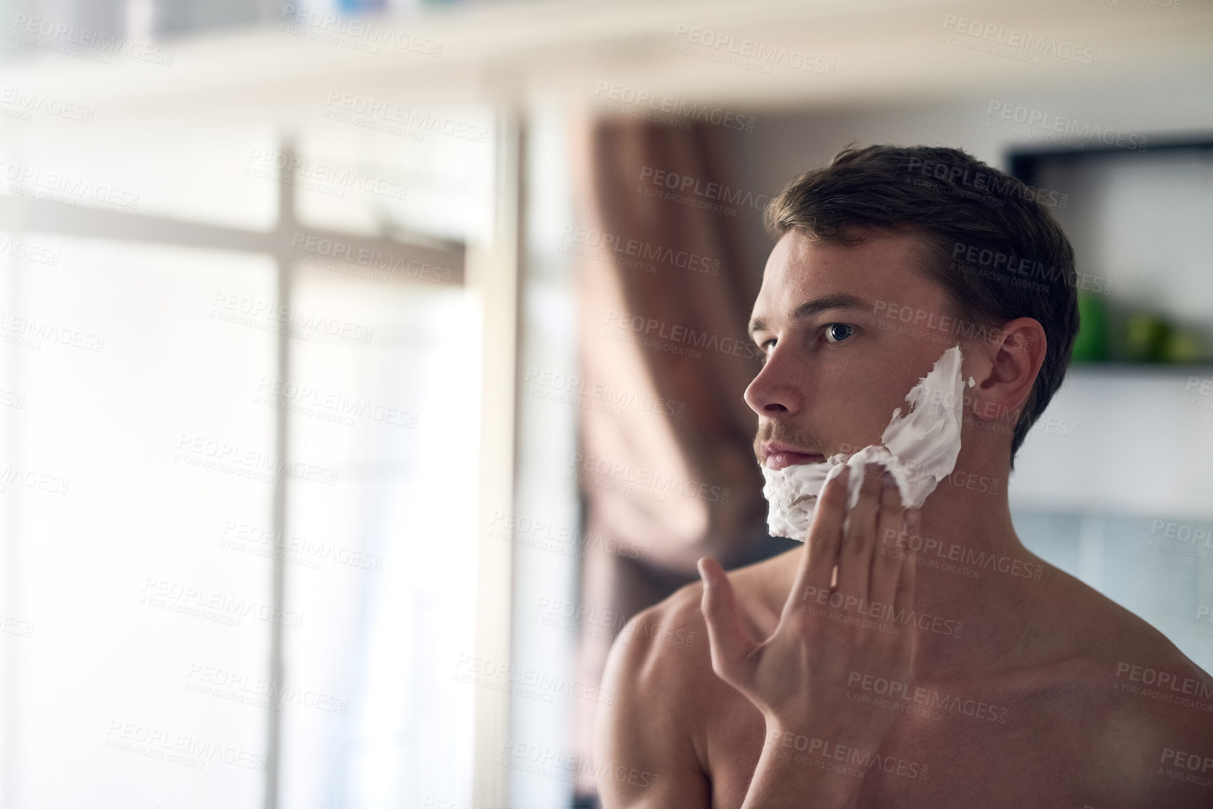 Buy stock photo Shot of a young man shaving his beard while looking at himself in the mirror