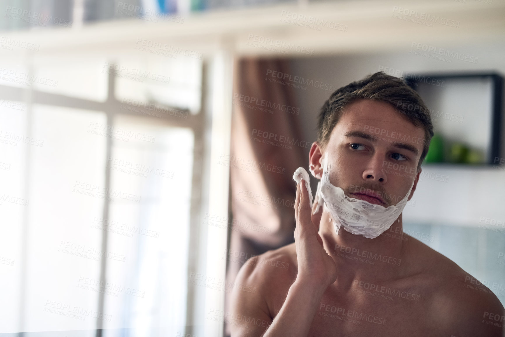 Buy stock photo Shot of a young man shaving his beard while looking at himself in the mirror