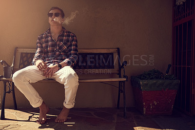 Buy stock photo Shot of a young man sitting on a bench outside and smoking a cigarette