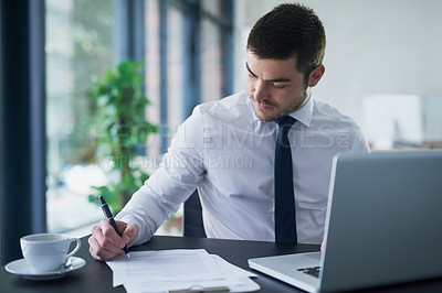 Buy stock photo Shot of a young businessman writing down notes while working on a laptop in an office
