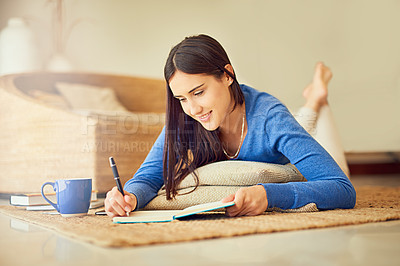 Buy stock photo Shot of a happy young woman making notes in her notebook while lying on her lounge floor