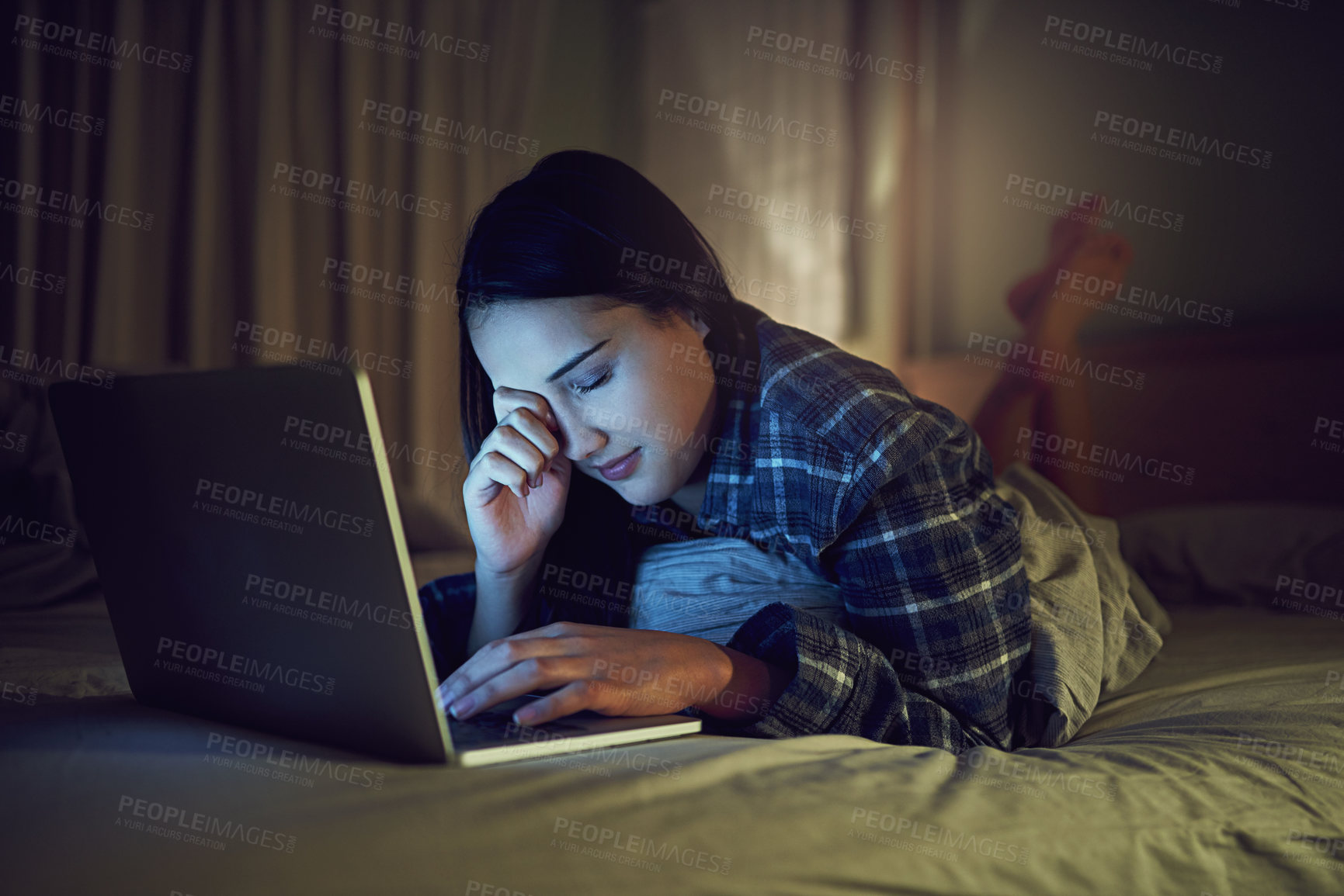 Buy stock photo Shot of a sleepy young woman using a laptop late at night while lying on her bed in her bedroom