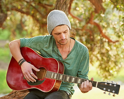 Buy stock photo Shot of a handsome young man sitting outside in a park playing guitar