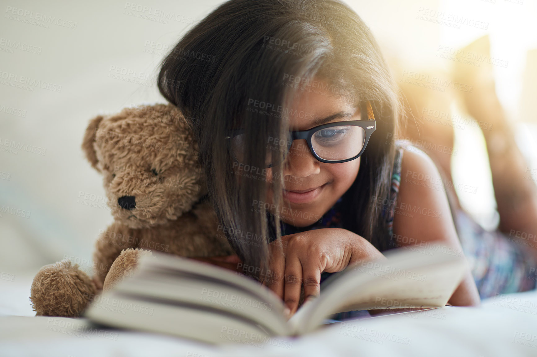 Buy stock photo Shot of a cute little girl reading a book in her bedroom with her teddybear by her side